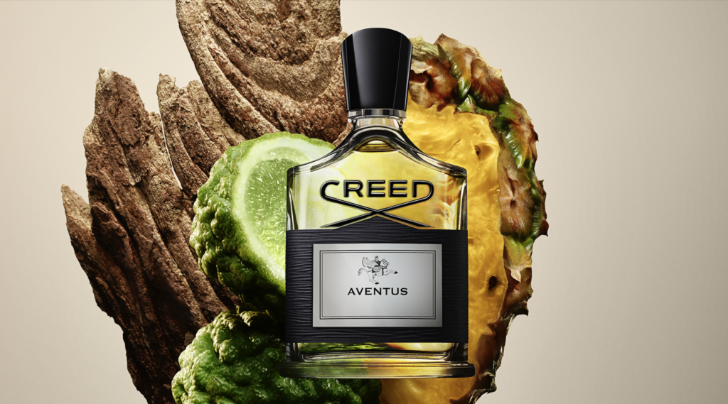 Aventus 3 - Petróleo y Energía Creed Aventus perfume bottle centered with pineapple and lime slices and rugged bark in the background.