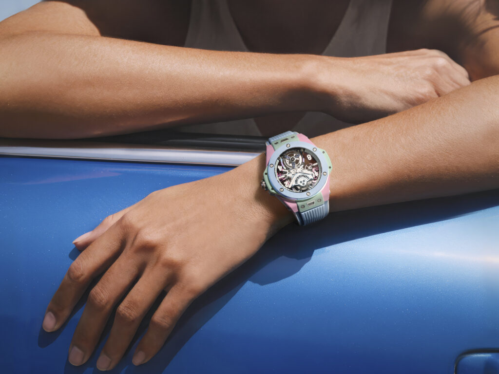 BIG BANG MULTI COLORED CERAMIC 44MM 2 - Petróleo y Energía Close-up of a forearm with a pastel multicolor wristwatch resting on a blue surface. The watch has a skeleton dial showing gears and pink/teal accents in the band.