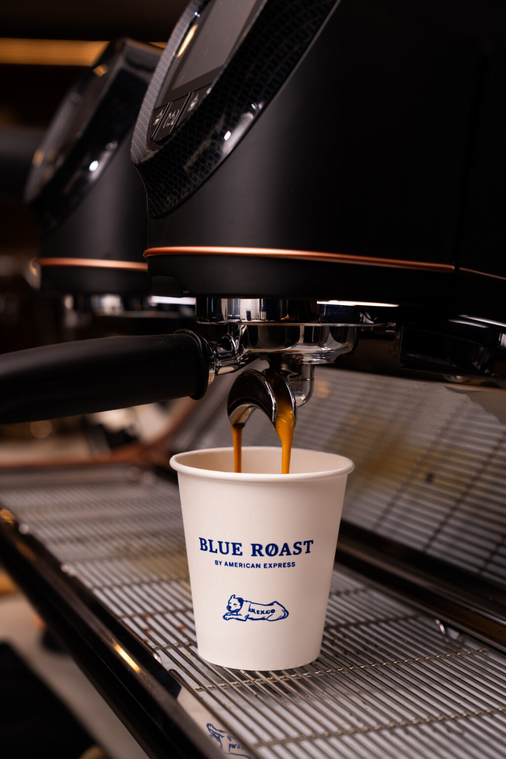 BLUE-ROAST_3003 1 - Petróleo y Energía Espresso machine pours dark coffee into a white cup labeled Blue Roast as it sits on a metal grate.