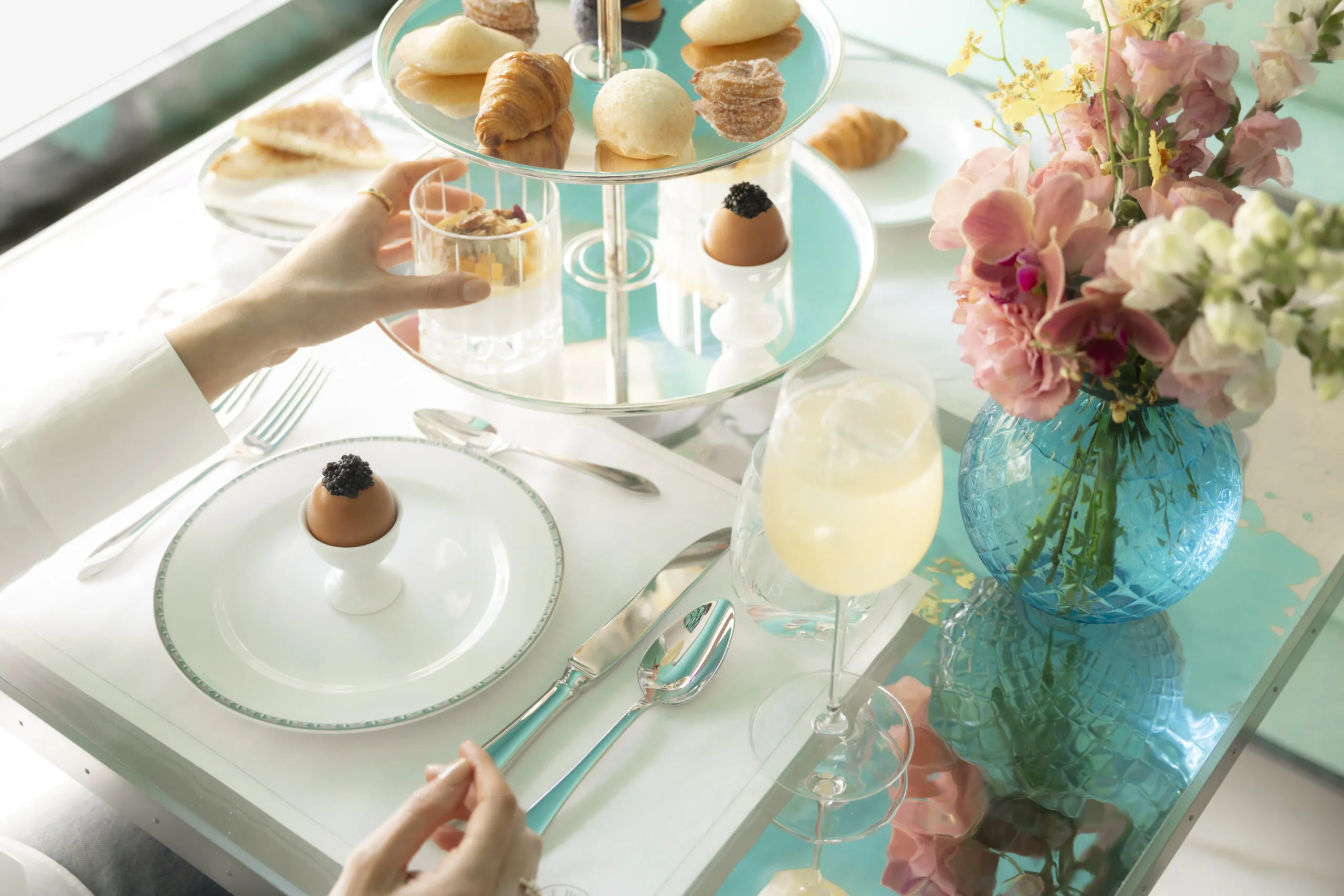 A hand reaching for a dessert on a tiered tray at a bright table with pastries, eggs, and a vase of pink flowers nearby.