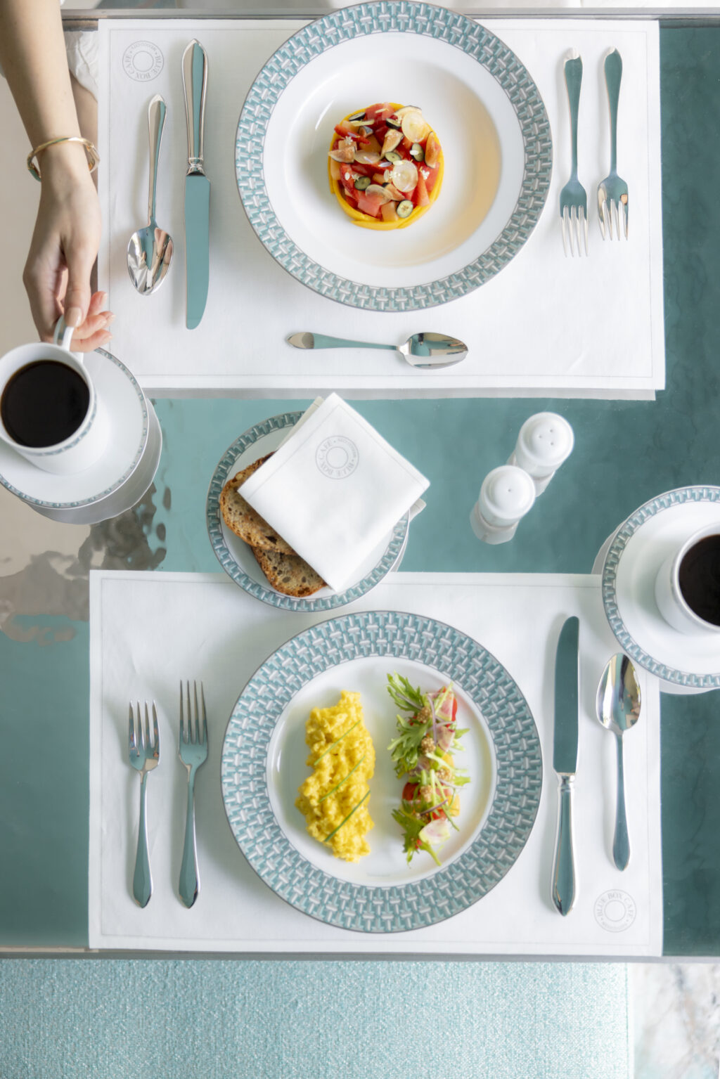 BLUEBOXCAFETIFFANY3727 - Petróleo y Energía Top-down view of a turquoise and white dining setting with patterned plates and silverware; a small fruit salad sits on the top plate, a hand pours coffee from a cup on the left, and a plate with scrambled eggs and greens is at the bottom.
