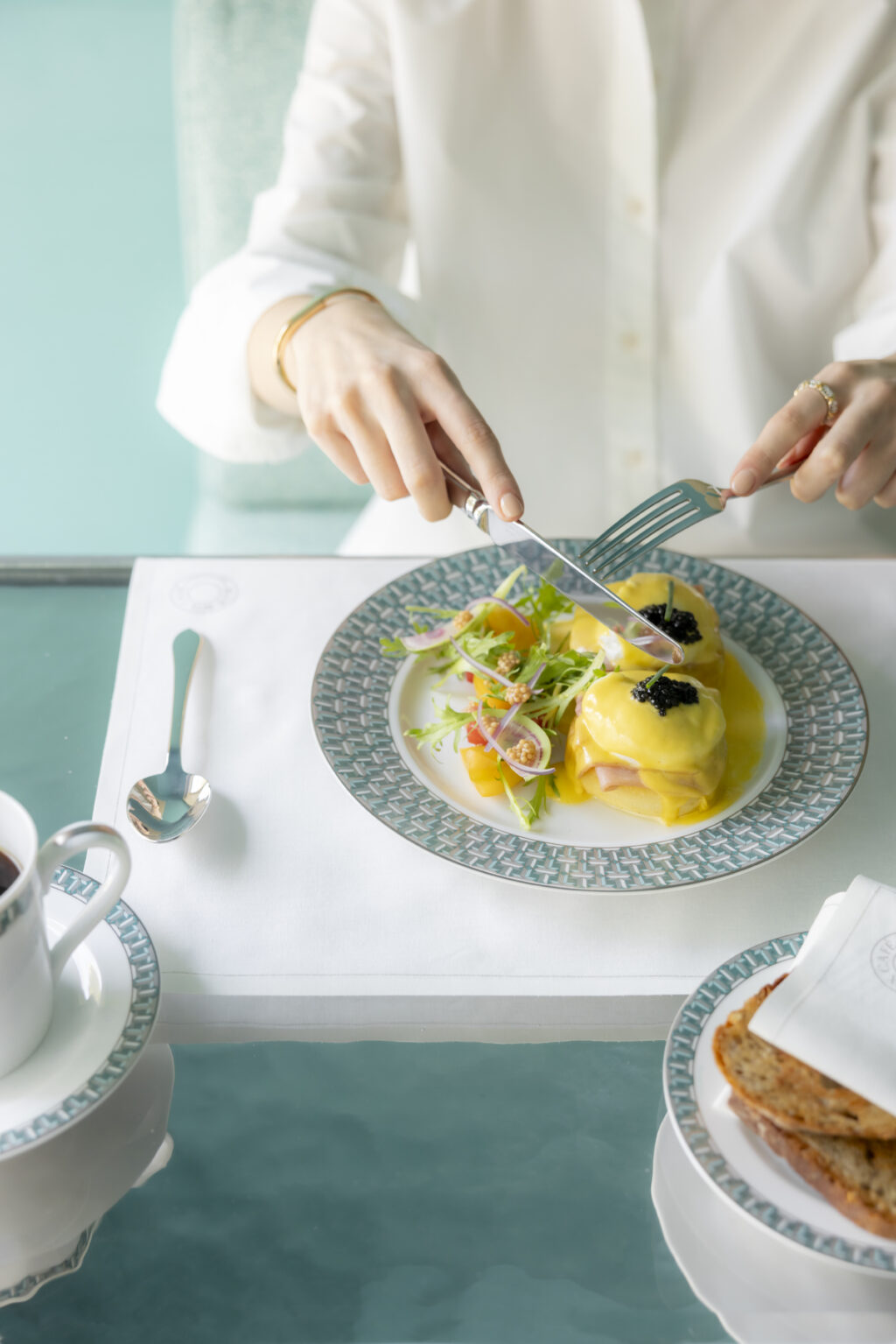 BLUEBOXCAFETIFFANY3776 - Petróleo y Energía Person in a white shirt uses a knife and fork to cut a plated dish with yellow sauce and greens on a patterned plate at a turquoise table.