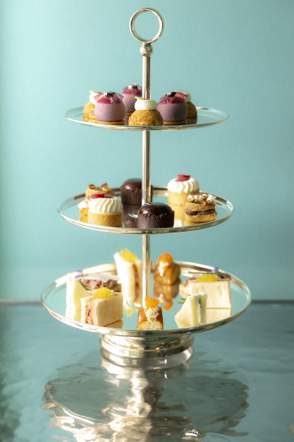 BLUEBOXCAFETIFFANY4802 - Petróleo y Energía Three-tier silver dessert stand with colorful pastries and sandwiches against a turquoise background.