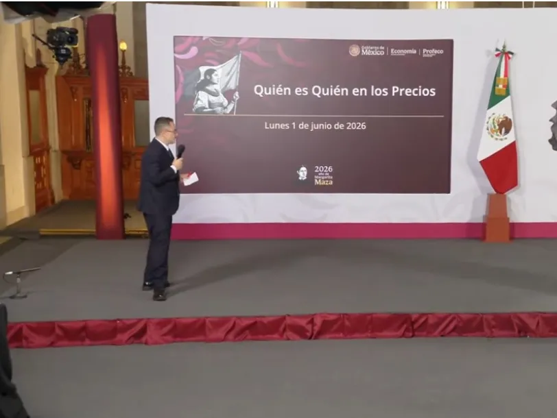 Presentador de traje hablando con micrófono en un escenario frente a una pantalla que anuncia 'Quién es Quién en los Precios'.