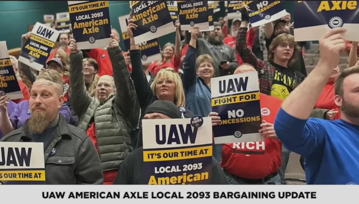 Crowd of union protesters holding UAW and no-concessions signs at a bargaining update event, with banner text at bottom reading 'UAW American Axle Local 2093 Bargaining Update'
