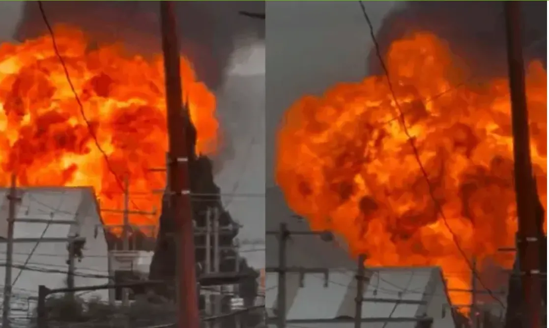 Two large orange explosion plumes rising over an industrial area with utility poles and wires in the foreground.