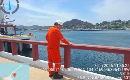 Worker in orange safety suit and white hard hat leaning on a railing at a dock, overlooking blue water and distant hills/ town.