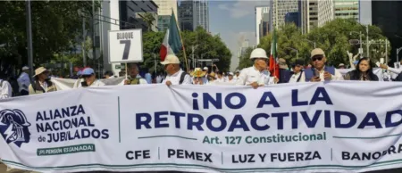 Protest march on a city street with a large banner reading '¡NO A LA RETROACTIVIDAD' and 'Art. 127 Constitucional', people holding the banner and Mexican flags, sign reading 'BLOQUE 7' visible in the crowd.