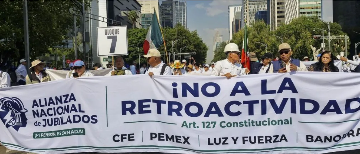 Protest march on a city street with a large banner reading '¡NO A LA RETROACTIVIDAD' and 'Art. 127 Constitucional', people holding the banner and Mexican flags, sign reading 'BLOQUE 7' visible in the crowd.
