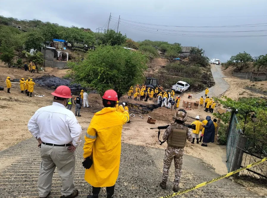 Emergency responders in yellow raincoats and red helmets stand around a muddy hillside disaster site with vehicles nearby.