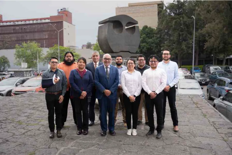 Group of professionals posing for a photo on a stone terrace with a large abstract sculpture behind them, in an urban area with cars in the background.
