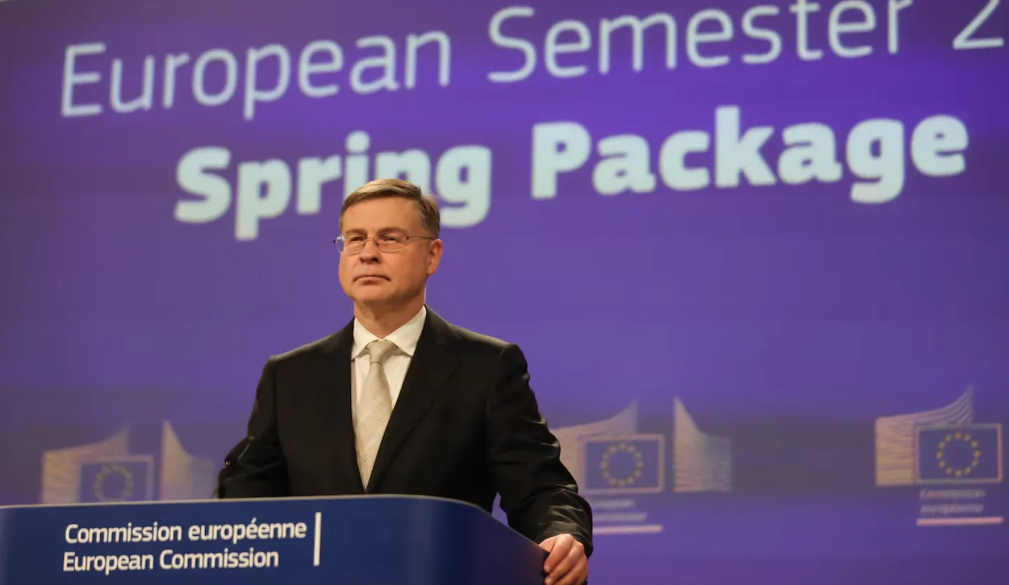 Man in a dark suit and tie delivering a speech at a European Commission podium, with a purple backdrop showing 'Spring Package' and EU symbols in the background.