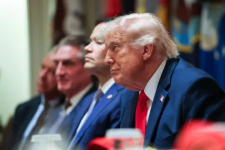 Side profile of a man with light hair in a navy suit and red tie, seated at a panel with other men in suits and flags in the background.