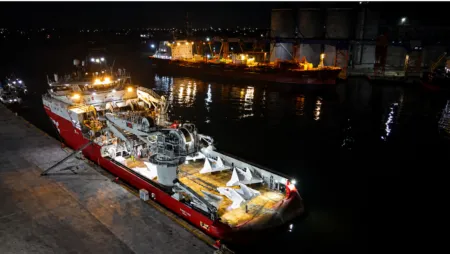 Nighttime port scene with a red and white offshore support vessel docked at a pier, deck machinery exposed and lit by work lights.