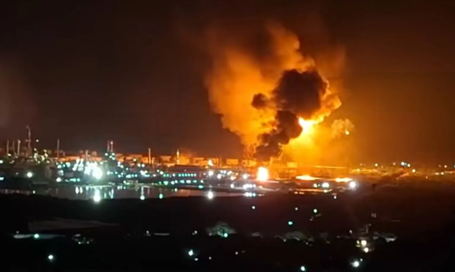 Nighttime industrial site on fire with large orange flames and thick black smoke rising above buildings, suggesting a refinery or plant.