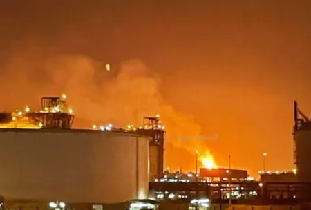 Night view of an oil refinery on fire, with large orange flames and thick smoke rising above industrial tanks and structures at night by a bright glow.
