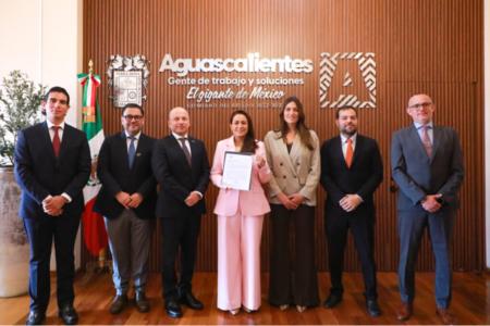 Seven professionals in formal attire pose in a government reception area; the central woman in pink holds a certificate, with the Aguascalientes seal and flag behind them.