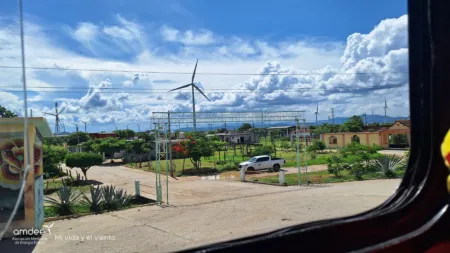 Wind turbines along a small town street under a bright blue sky with fluffy clouds; a white pickup parked on a dirt road in the foreground.