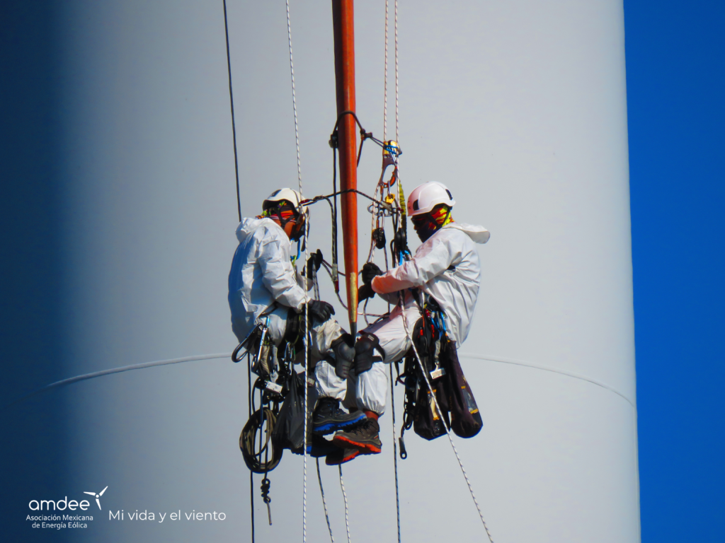 Copia de victor-abraham-trabajo-y-el-viento-260610121449_yiq6qEg2 - Petróleo y Energía Two workers in white protective suits and helmets climbing a wind turbine mast with ropes and harnesses, against a blue sky. (AMDEE branding visible)