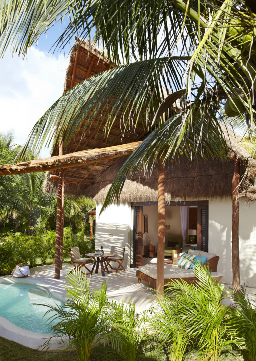 Copy of VRM20 - Petróleo y Energía Tropical resort scene with a thatched-roof bungalow, palm fronds overhead, and a turquoise pool with outdoor seating nearby.