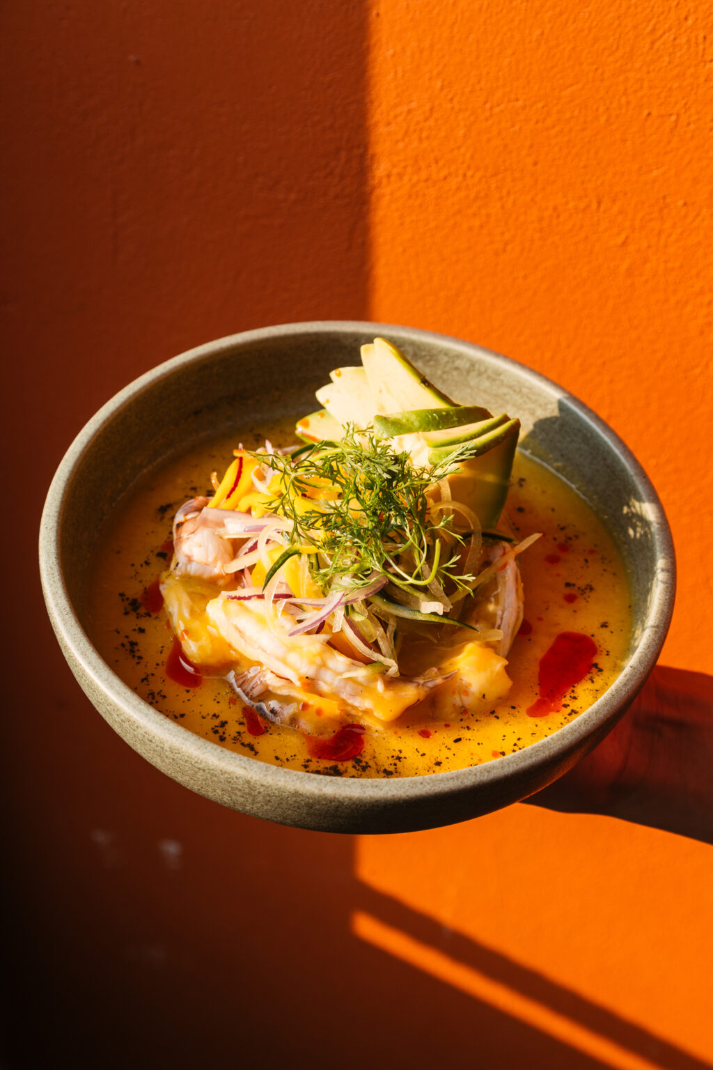 Copy of _MG_1339 - Petróleo y Energía Bowl of creamy yellow seafood soup topped with shrimp, herbs, and lime wedges on an orange background.