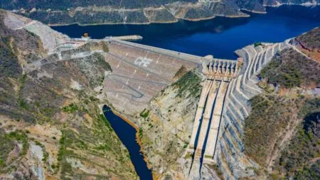 Aerial view of a large concrete dam with long spillways along a rocky canyon and a deep blue reservoir nearby.