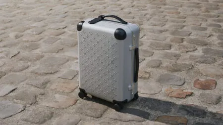 Silver Louis Vuitton hard-shell suitcase with embossed monogram pattern on a cobblestone pavement, upright on four wheels with a top handle visible.