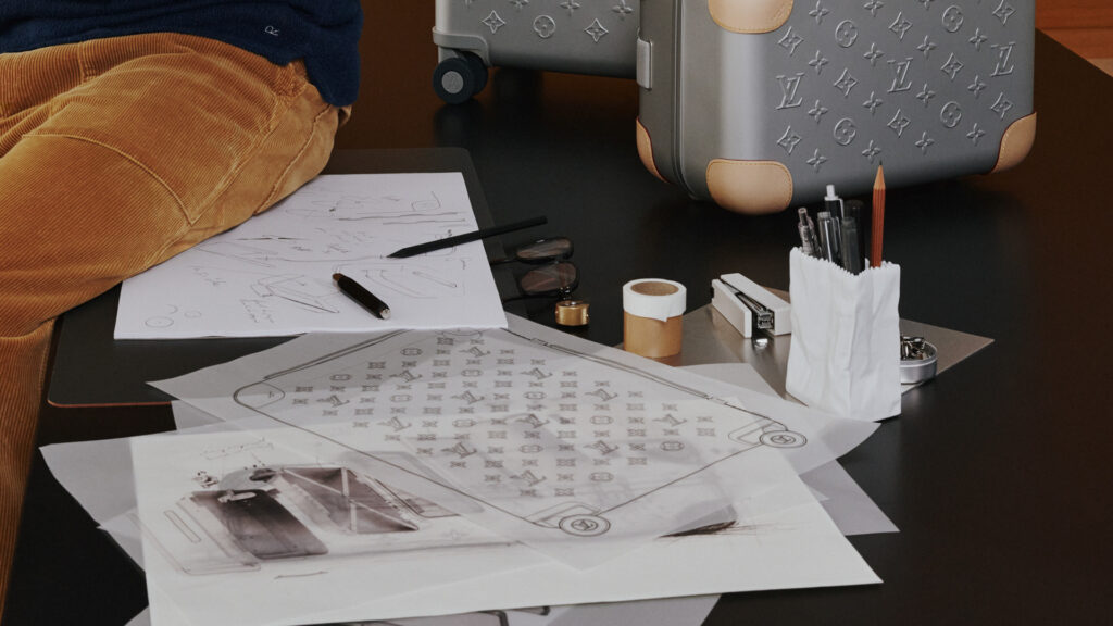 HORIZON_ALUMINUM_WORN_PICTURES_8_-_1920x1080 - Petróleo y Energía Studio desk with sketches, rolled papers, a Louis Vuitton patterned suitcase, and drafting supplies.