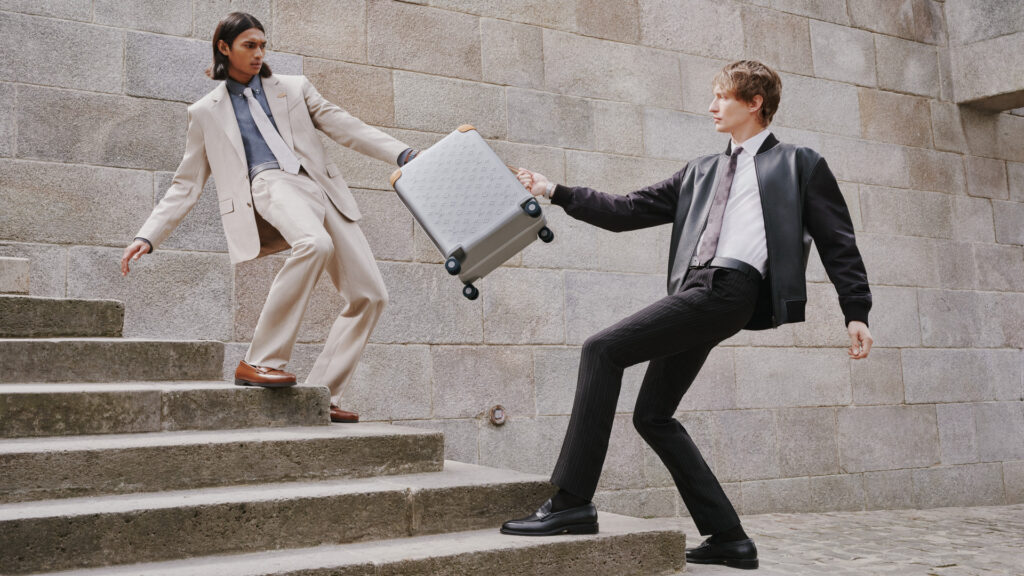 HORIZON_ALUMINUM_WORN_PICTURES_9_-_1920x1080 - Petróleo y Energía Two men in suits on stone stairs, one handing a rolling suitcase to the other midair in a dramatic scene behind a concrete wall.