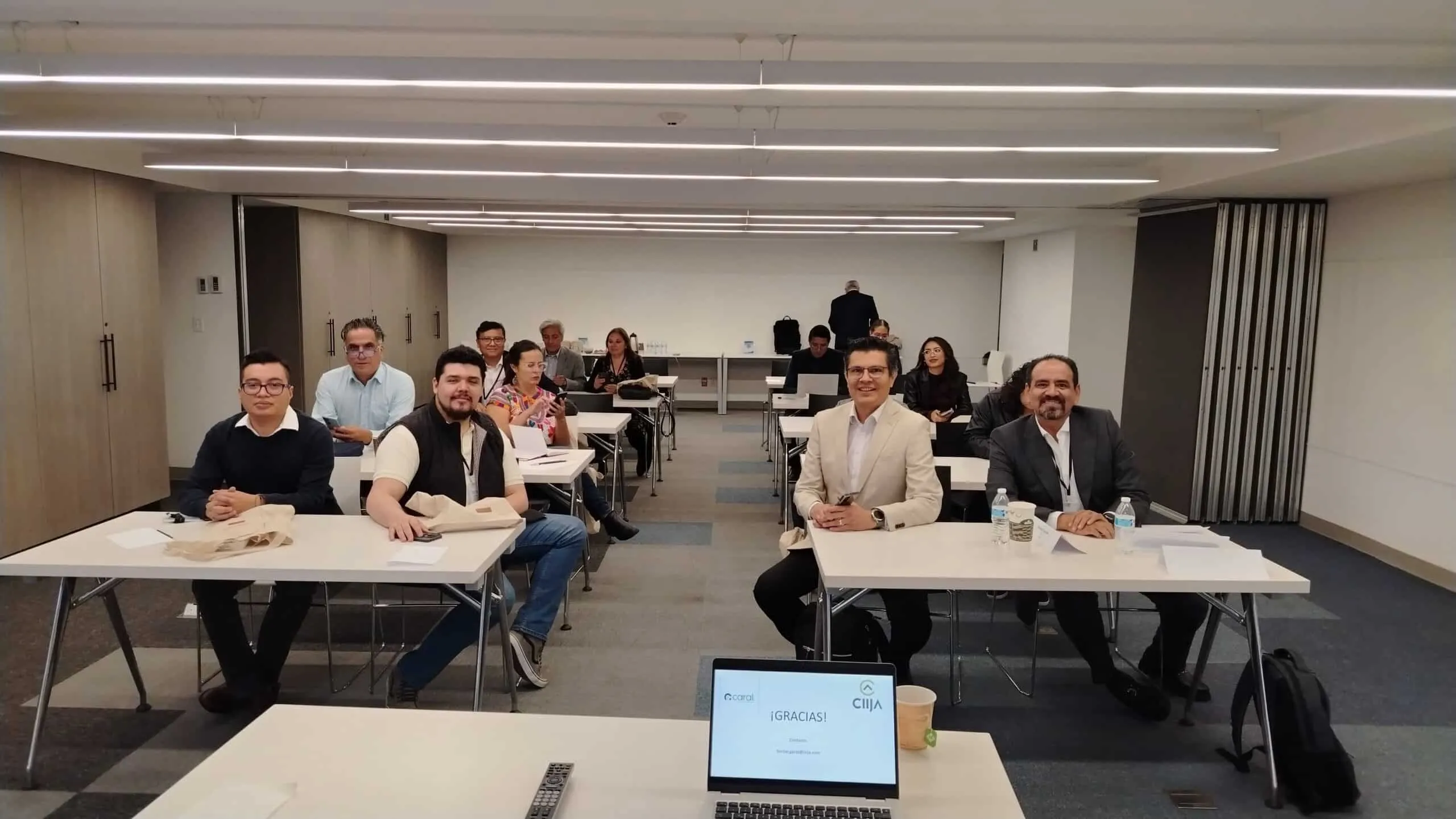 Group of professionals seated around tables in a conference room, facing the camera, with a laptop showing '¡GRACIAS!' in the foreground .