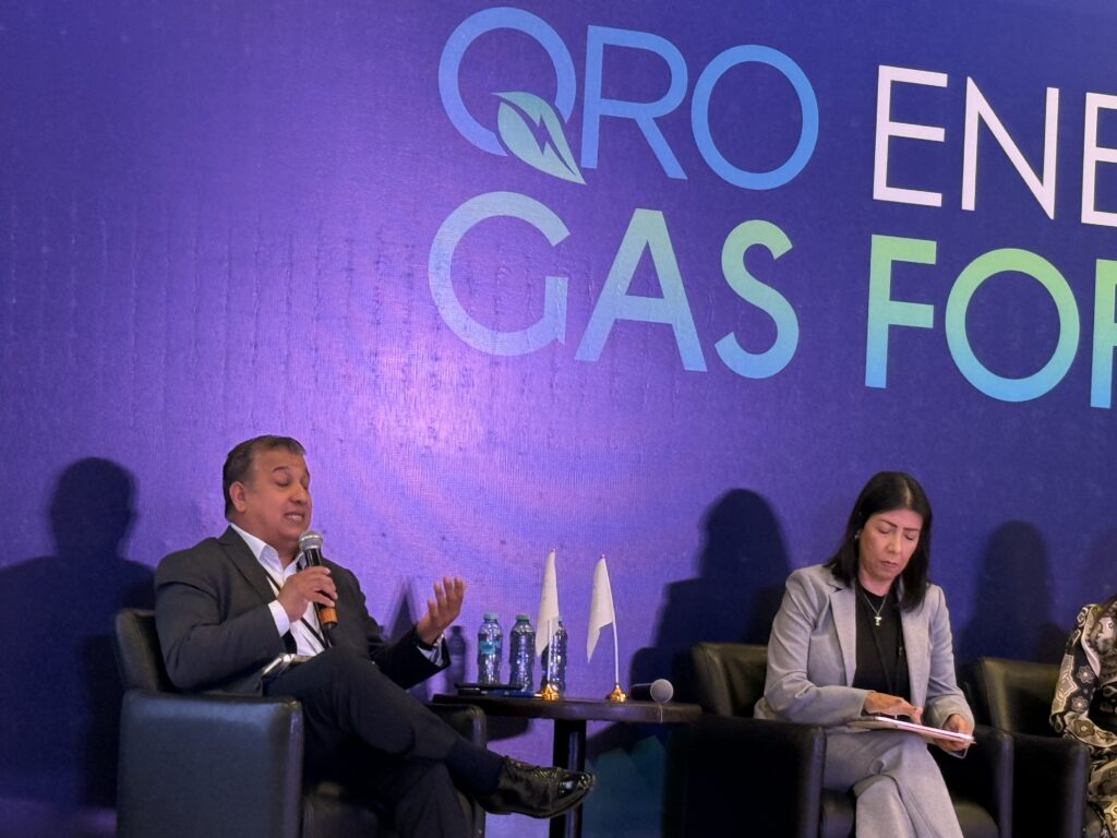 - Petróleo y Energía Two panelists sit on a stage with a man speaking into a microphone and a woman taking notes, water bottles on the table, purple backdrop text partly visible behind them.