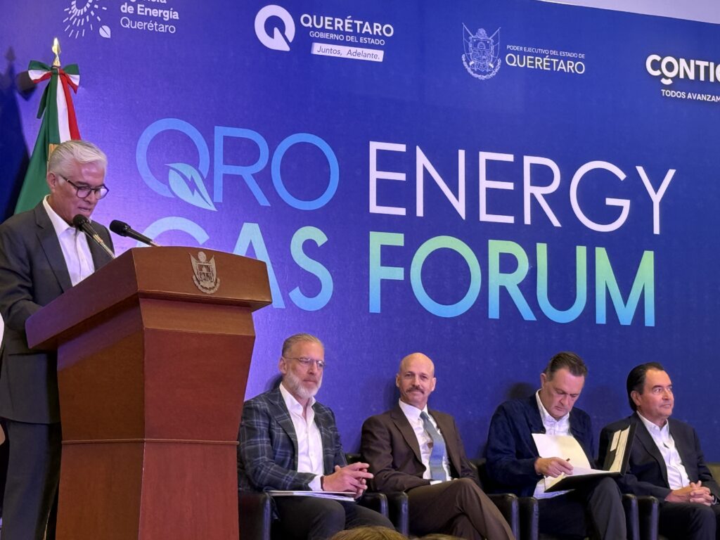 - Petróleo y Energía Man in a suit speaking at a wooden podium during a formal energy forum, with a blue backdrop and seated panelists behind him.