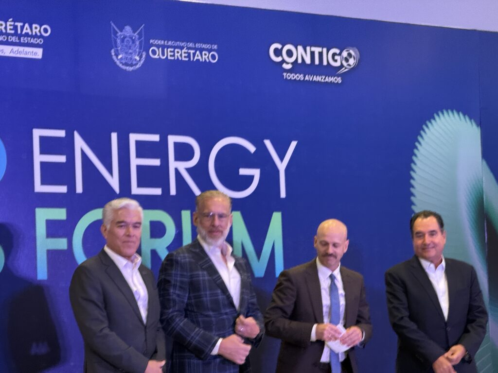 - Petróleo y Energía Five men in suits standing on a stage in front of a blue backdrop with ENERGY text; two in the foreground appear to be shaking hands while others stand nearby.