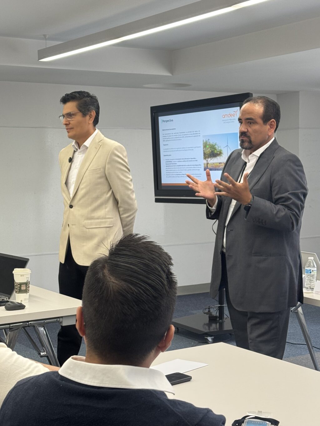 IMG_7129 - Petróleo y Energía Two men in business attire giving a presentation in a conference room with a large screen showing slides behind them