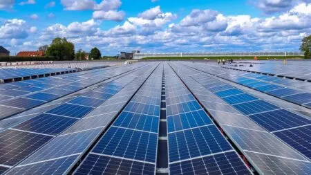 A vast solar farm with long rows of blue panels under a bright, partly cloudy sky; workers in high-visibility vests in the distance.