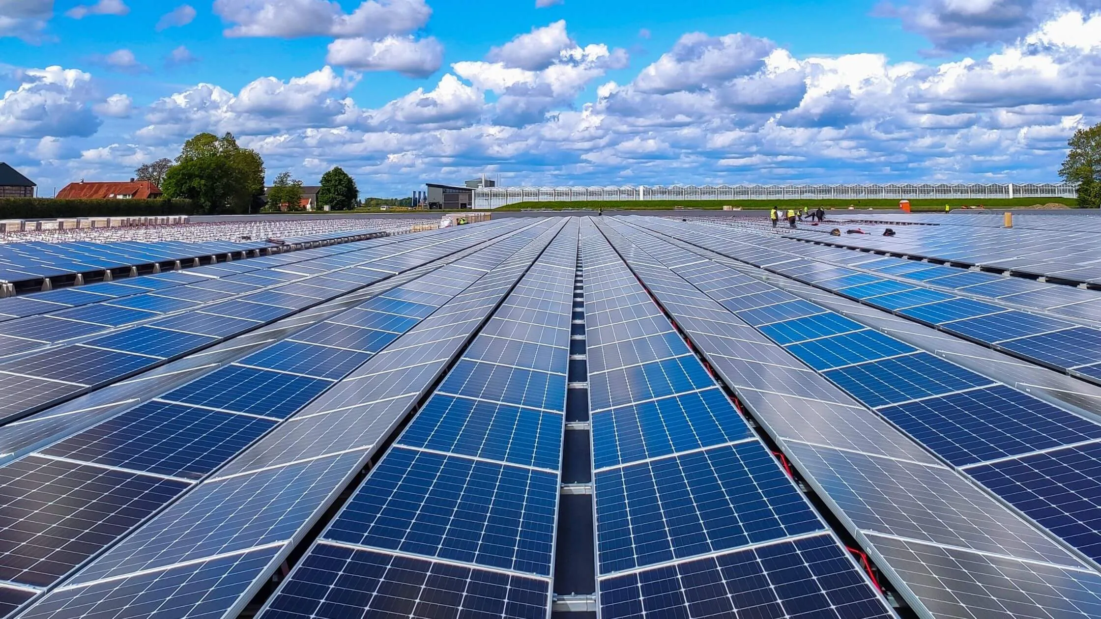 A vast solar farm with long rows of blue panels under a bright, partly cloudy sky; workers in high-visibility vests in the distance.