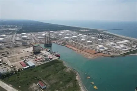 Aerial view of an industrial port or refinery complex with storage tanks, cranes, and docks along a coastline.