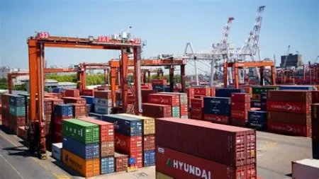 Shipping container yard with large orange gantry cranes and stacked colorful containers under a clear blue sky.