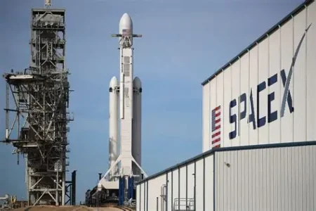SpaceX Falcon rocket on the launch pad between a towering service structure and a hangar labeled SPACEX under a clear blue sky.