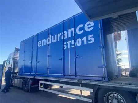 Blue cargo trailer on a truck with 'Endurance' and 'STE5015' visible; a person stands near the cab at a loading area.