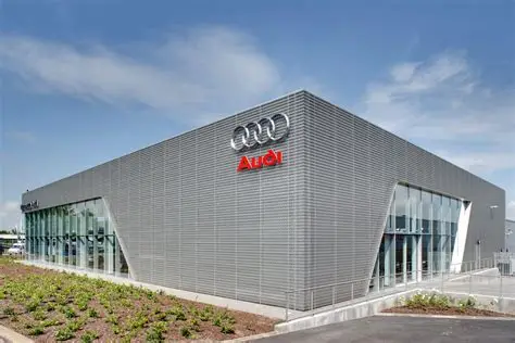 Audi dealership building with metal panel exterior and glass showroom walls, displaying the Audi logo on the facade.