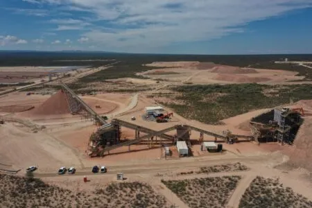 Open-pit mining site with conveyor belts, large ore piles, and desert landscape under a blue sky; several trucks parked near the yard.