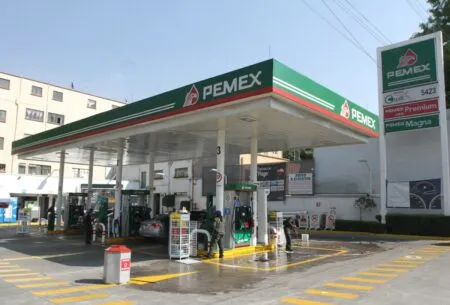Pemex gas station with a green canopy and multiple pumps; workers refuel cars on a wet forecourt under a clear blue sky.