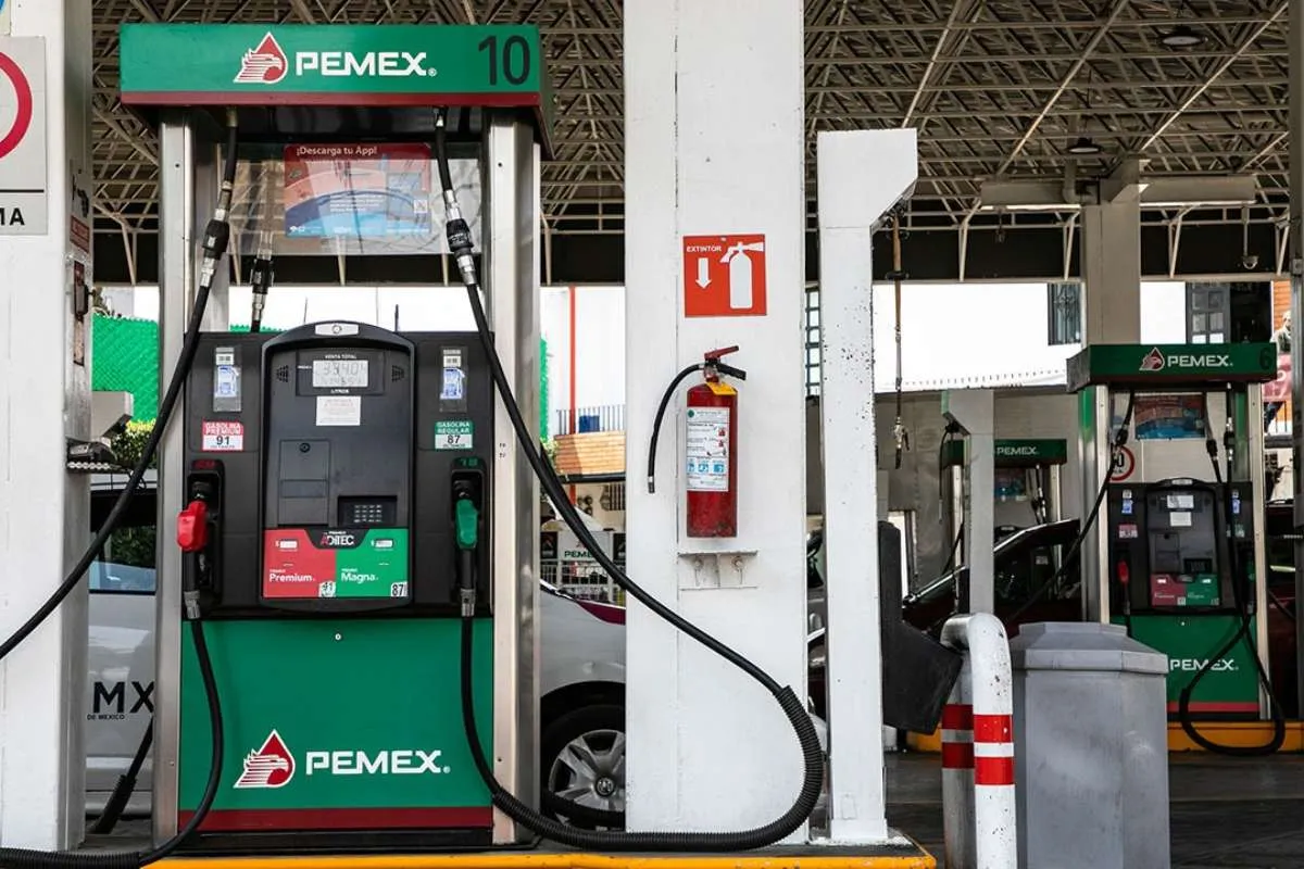 Gasoline pumps at a PEMEX station with green branding, hose lines, and a fire extinguisher mounted on a white pillar in the foreground.