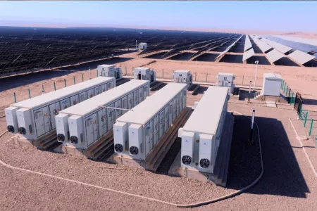 Desert solar farm with long rows of solar panels and multiple large energy storage containers in the foreground connected by cables and fenced area.