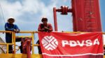 Two oil workers in hard hats lean on a yellow railing beside red piping, a PDVSA banner dangling in front of them on a sunny day