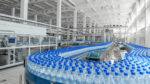 Industrial bottling plant with a circular conveyor of water bottles with blue caps, under bright white lighting and stainless machinery.