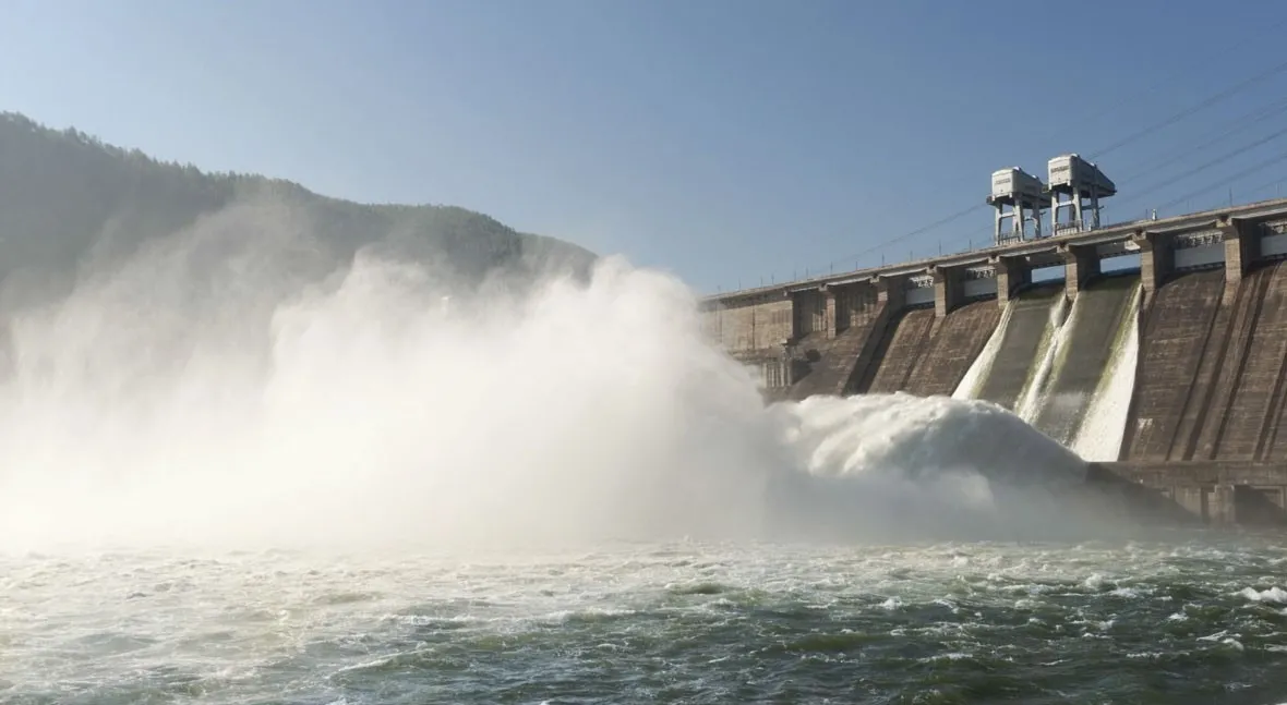 Dam spillway releasing a powerful flow of water into a river with mist rising and hills in the background.