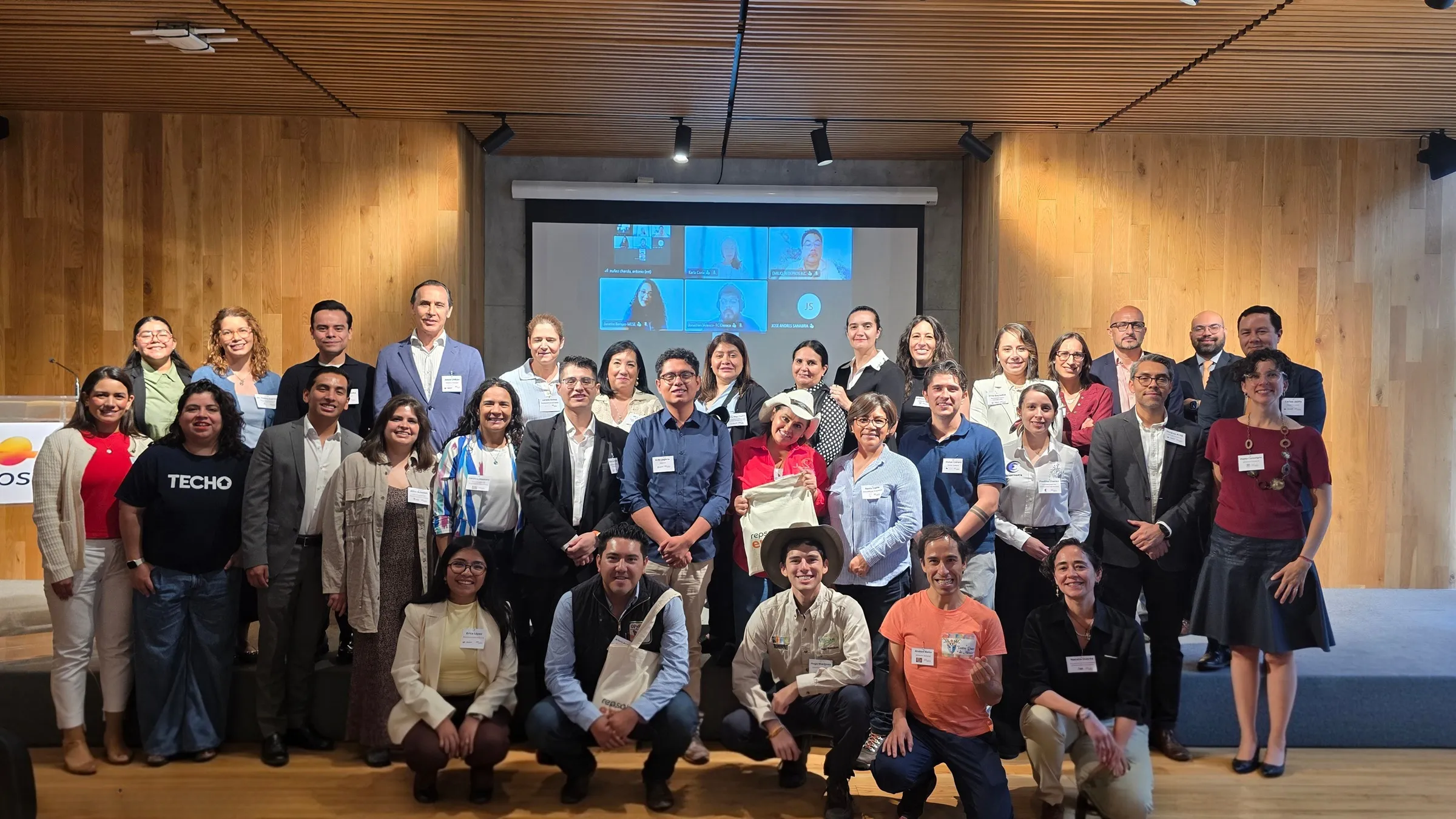 Large group of diverse attendees posing on a wood-paneled conference stage, with a projection screen showing a video call in the background.