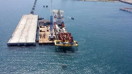 Aerial view of a harbor with a long concrete floating pier, a yellow workboat, and industrial equipment on a platform at sea.